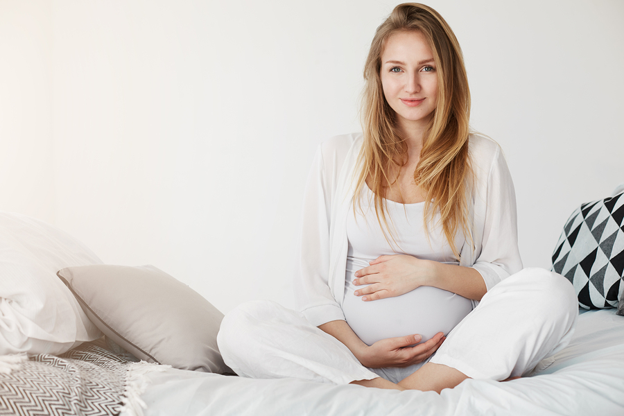 Para ilustrar as vantagens do colágeno para grávidas, uma mulher gestante e de aparência jovem está sentada sobre uma cama segura a barriga e encara a câmera.