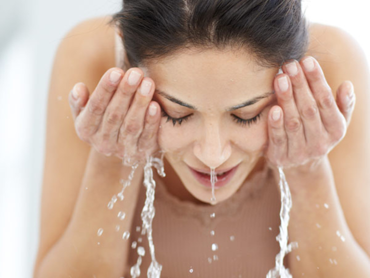 Mulher inclinada para frente lava o rosto. Escorre água de suas mãos e seu nariz.
