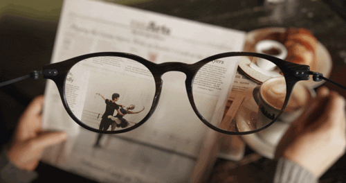 Através das lentes de um óculos, vemos um jornal e uma xícara de café.