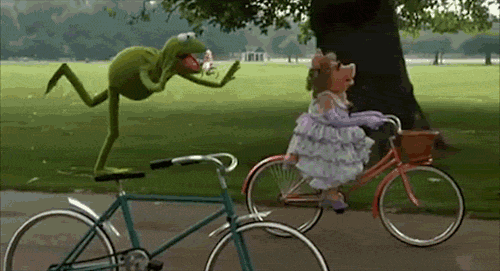 Caco, fantoche de um Sapo que é personagem do seriado Muppets, equilibra-se com um pé só de pé no banco de uma bicicleta. Ao seu lado, está Miss Piggy, fantoche de um pouco vestido de mulher que interpreta outro personagem. Ela pedala em sua própria bicicleta. Os dois estão num parque árvores, grama verde e um lago ao fundo.