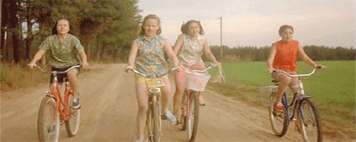 Quatro meninas andam de bicicleta por uma rua de terra.