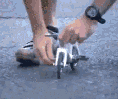 Homem pedala numa bicicleta em miniatura, do tamanho de seus sapatos.
