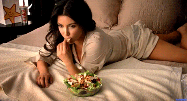Mulher deitada de bruços numa cama de casal come uma salada.